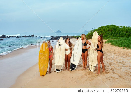 Group of diverse female surfers stand with boards on beach. Women smile, talk, enjoy surf culture by ocean. Swimsuits, tropical waves, sandy coast. Sport, friendship, adventure on sunny day. 122079383