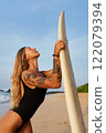 Tattooed young woman surfer holds surfboard on sunny beach. Blonde hair, swimsuit, ocean waves. Fitness, surf lifestyle, travel, adventure. Sun tanned skin, coastal vibes, outdoors exercise. 122079394