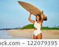 Asian female surfer carries surfboard on sunny beach. Smiling woman in swimsuit walks on sandy shore after surf. Coastal landscape with blue sky, green plants. Outdoor water sports, summer. 122079395