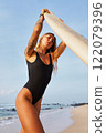 Woman poses on sandy beach with surfboard. Tattooed female in swimsuit enjoys summer by ocean. Surfing gear, blue sky, coastal landscape. Fitness, lifestyle, seaside adventure. 122079396