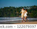 Romantic couple walk on sandy beach, man and woman hold tropical drinks, stroll by sea during sunset. Barefoot honeymooners enjoy ocean waves, relaxed vacation. Love, travel, summer concept captured. 122079397