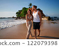 Couple walks barefoot on sandy beach by ocean at sunset. Man and woman enjoy romantic stroll near waves. Romantic getaway, lovers beside sea. Holding hands, serene, peaceful beach walk, travel couple. 122079398