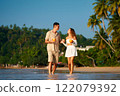 Couple stroll barefoot on sandy beach, holding coconuts, sipping drinks, enjoying sunset. Casual date, tropical vacation vibe. Smiles, flirty conversation, ocean backdrop, intimate walk together. 122079392