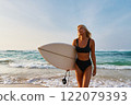 Blonde woman in bikini holds surfboard on sunny beach. Smiling female walks by ocean, enjoys summer, beach vibes, surf lessons. Active lifestyle at sea, leisure. Young surfer in swimwear. 122079393