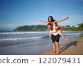 Man carries woman piggyback on sunny beach. Couple enjoys playful time by the sea. Smiling female spreads joy, freedom. Casual vacation, fun, summer travel, leisure activity at coastal landscape. 122079431