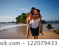 Happy couple enjoys beach getaway. Man carries woman piggyback by ocean. Playful pair laugh in sunlight, sandy shore. Casual dating, fun moments. Romantic walk, sea backdrop, holiday vibes. 122079433