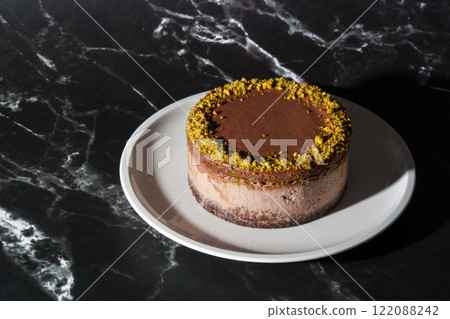 Dubai chocolate cheesecake with pistachio paste and kataifi filling covered in chocolate ganache on white plate. Black marble background with copy space. Indulgence, gourmet desserts, art of culinary 122088242