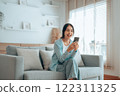 Smiling pretty young Asian woman sitting on cozy couch, using funny mobile apps in living room 122311325