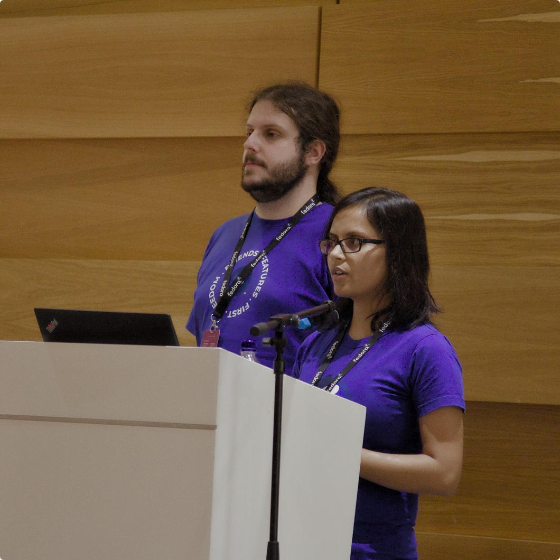 Two people weaing purple shirts standing behind a podium with a laptop presenting at a conference