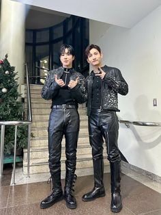 two men standing next to each other in front of a christmas tree and stairs wearing black leather pants