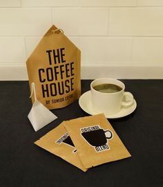 a cup of coffee sitting on top of a table next to two bags with the words the coffee house written on them