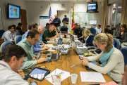 a group of people sitting around a wooden table working on laptops and papers in front of them