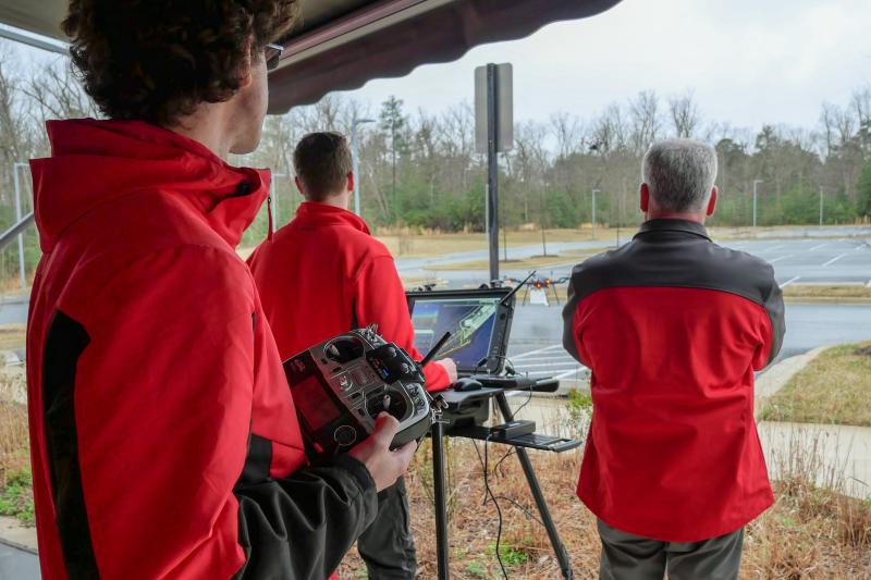 Three pilots watch drone fly, one holds another drone, one stands at computer