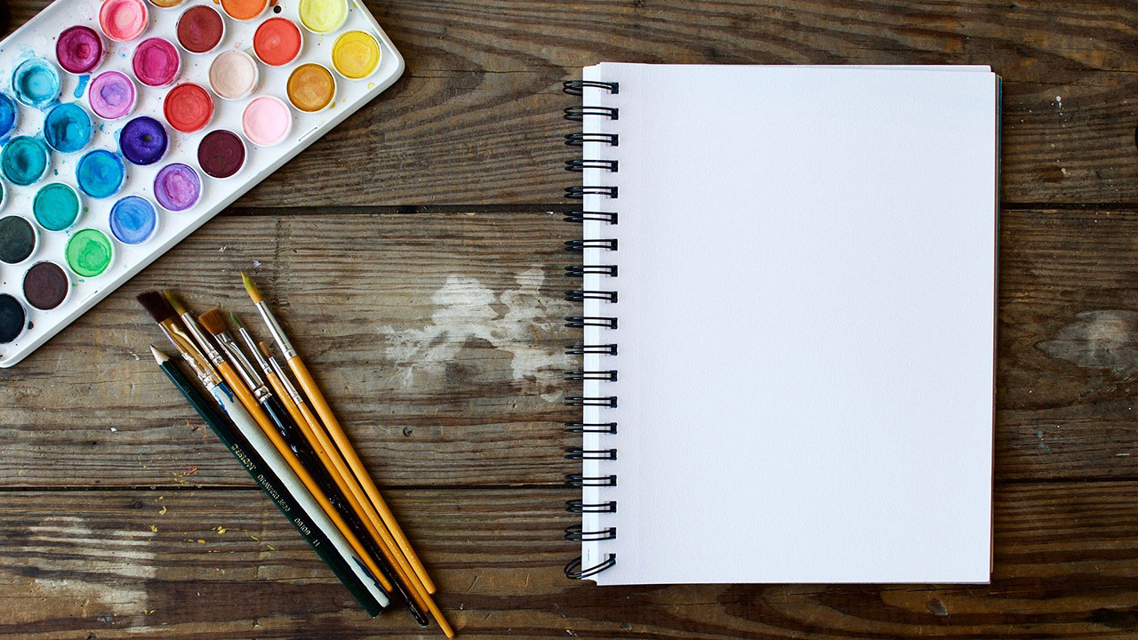 Coloured paint set and brushes beside a white paper notepad