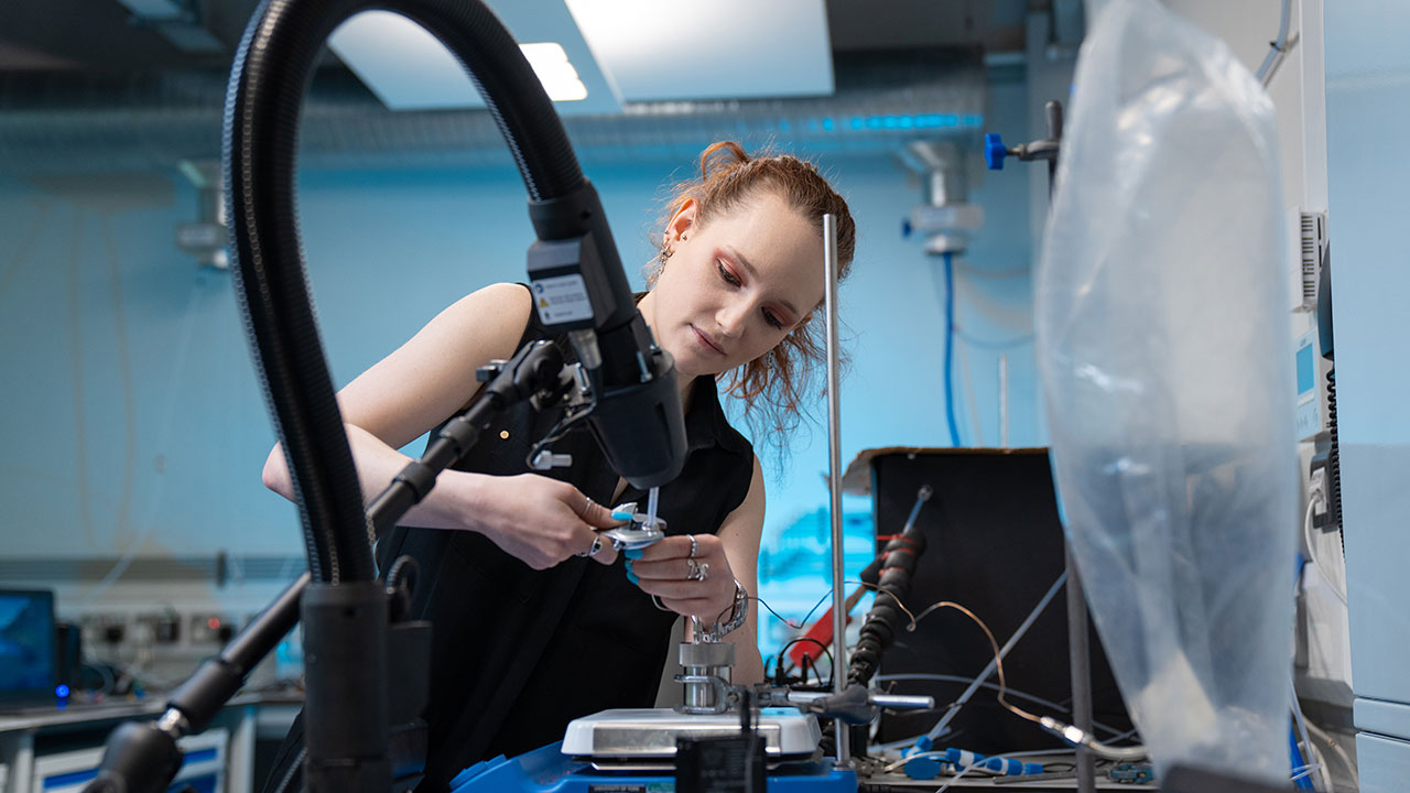 Person leans over laboratory desk, holding small wrench and gas inlet tube