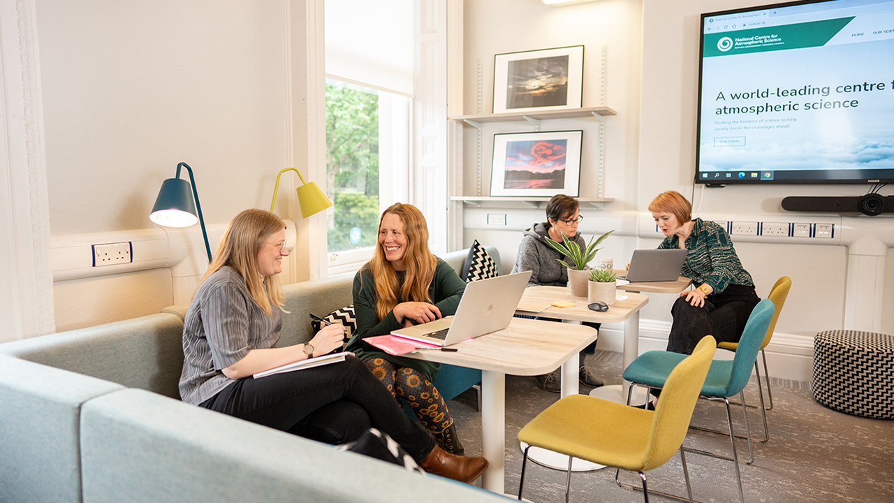 A group of four people chatting and looking at laptops and notepads in a communal workspace