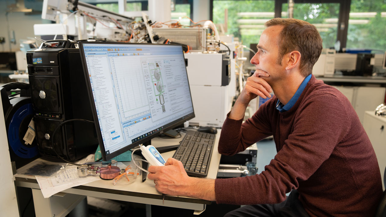 A male scientist looks at computer screens with scientific information on them. They are sat in a laboratory.