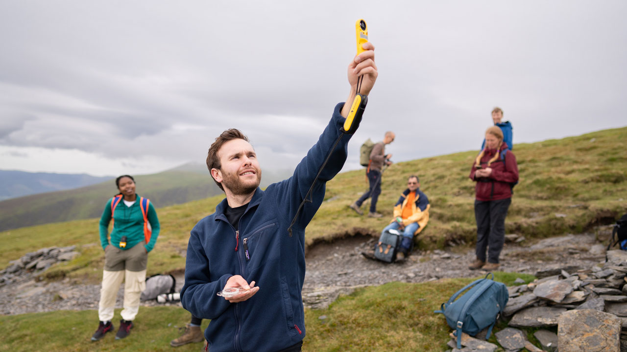 Person wearing a blue fleece holding a yellow weather monitoring instrument in the air. In their other hand is a compass. Green grass, hills and grey clouds in front of them.