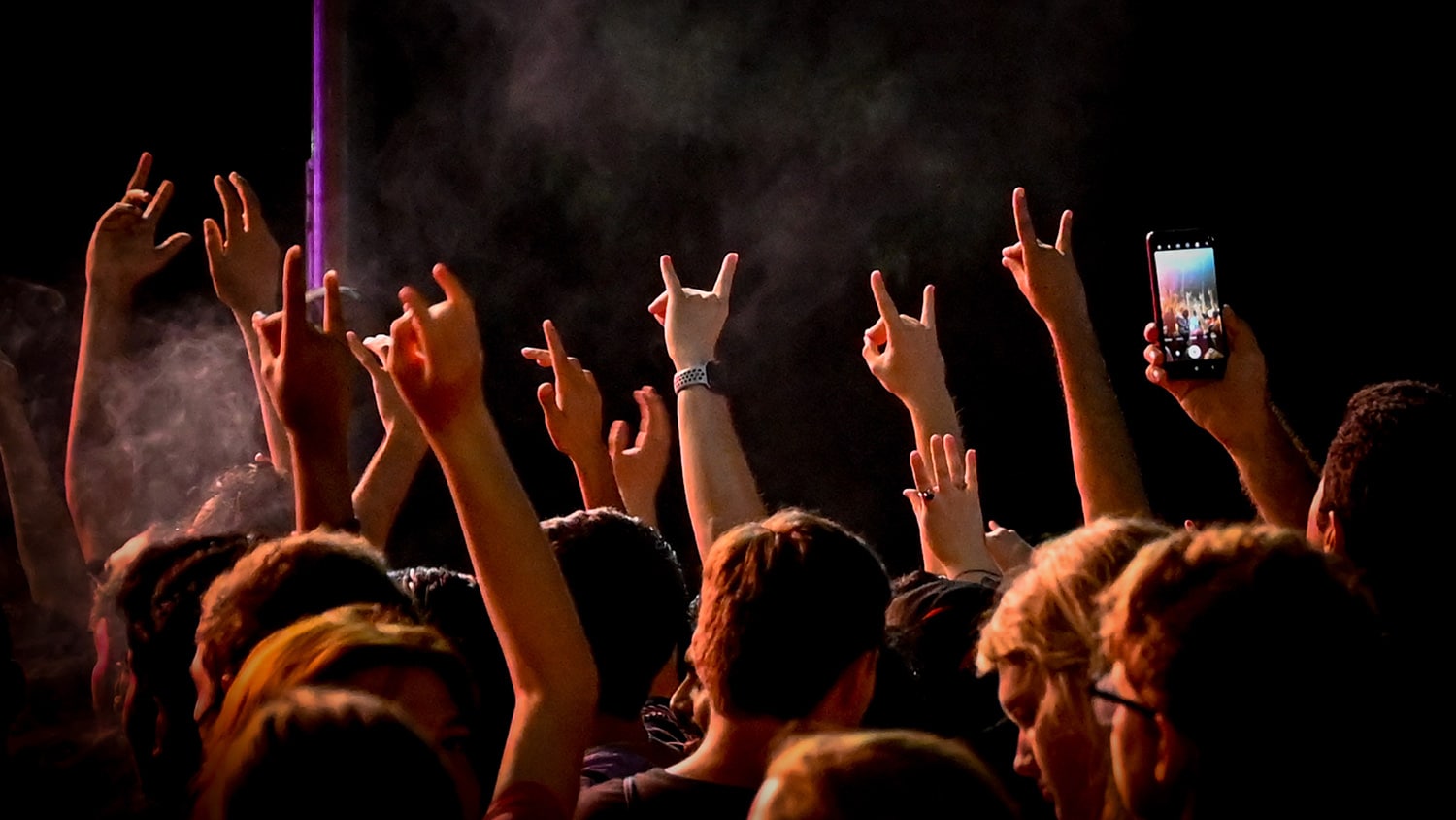 A large crowd, shown from behind, holds up the wolfie hand gesture during a nighttime concert.