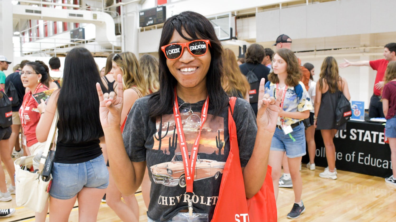 A student poses with both hands making wolfie gestures, wearing sunglasses that say "I Choose Education" on the lenses, in an auditorium full of people and information tables.