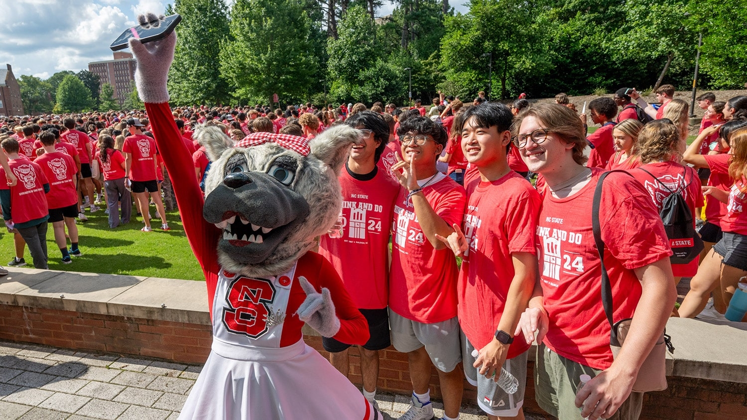 Four students in matching NC State T-shirts pose for a selfie with Ms. Wuf outdoors before taking their class photo during convocation.