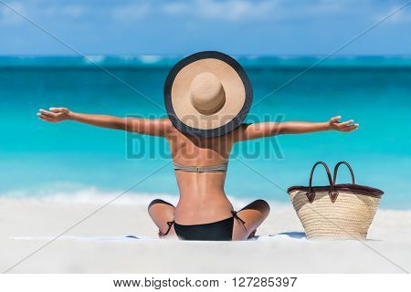 Summer vacation happy carefree joyful bikini woman arms outstretched in happiness enjoying tropical beach destination. Holiday girl sitting with sun hat relaxing from behind on Caribbean vacation.