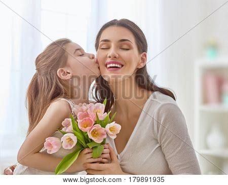 Happy mother's day! Child daughter congratulates mom and gives her flowers tulips. Mum and girl smiling and hugging. Family holiday and togetherness.