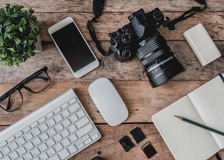 Camera, writing, and computer equipment are laid out neatly and evenly spaced out on a wooden desk. A fake plant adds some colour in the top left corner. 